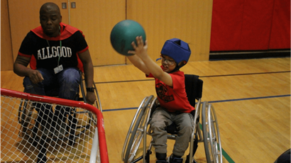  A student in a wheelchair lifts a ball over his head aiming for a net in a school gymnasium. 