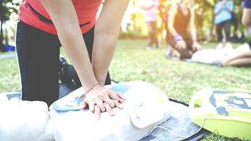 Woman in a CPR class in a park interlocks fingers and practices chest compressions on a mannequin that has an AED applied.
