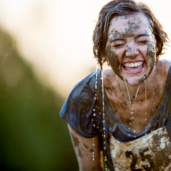 A smiling woman with mud on her face wears a race bib.