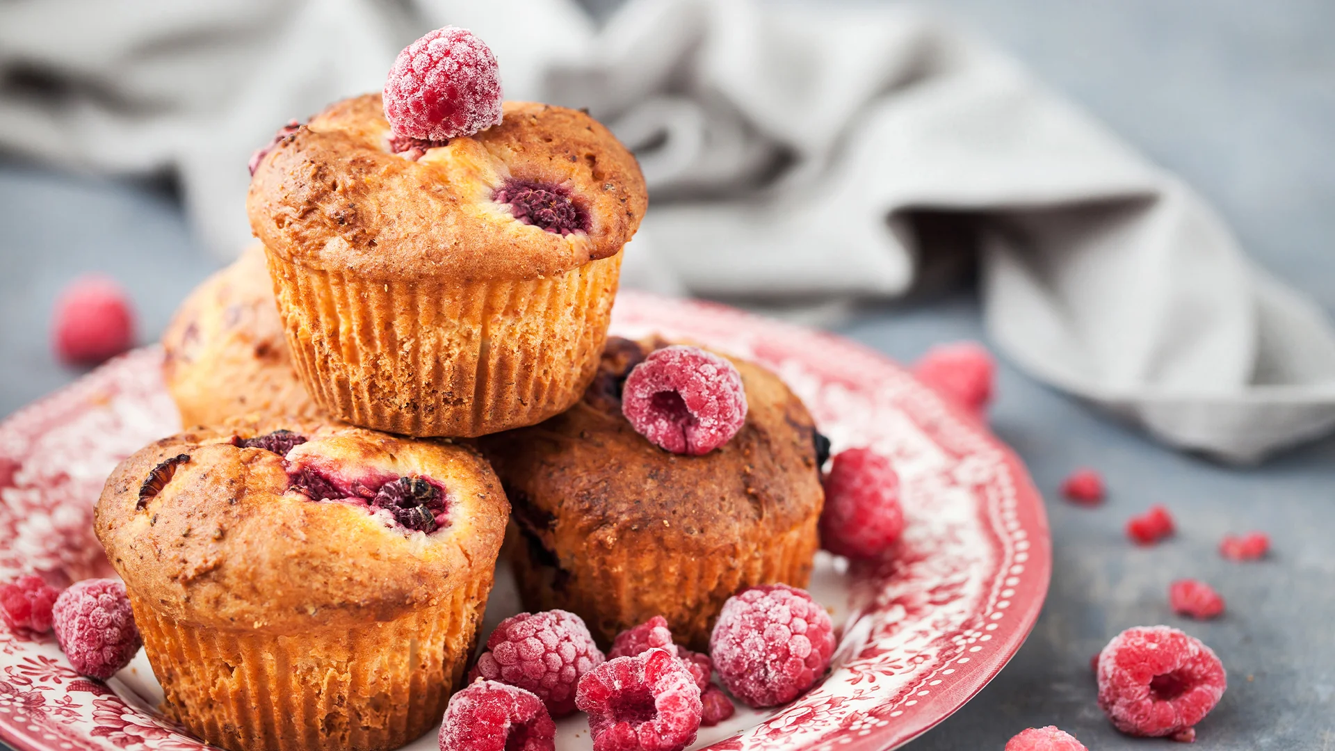 Four raspberry muffins on a dish garnished with frozen raspberries. 